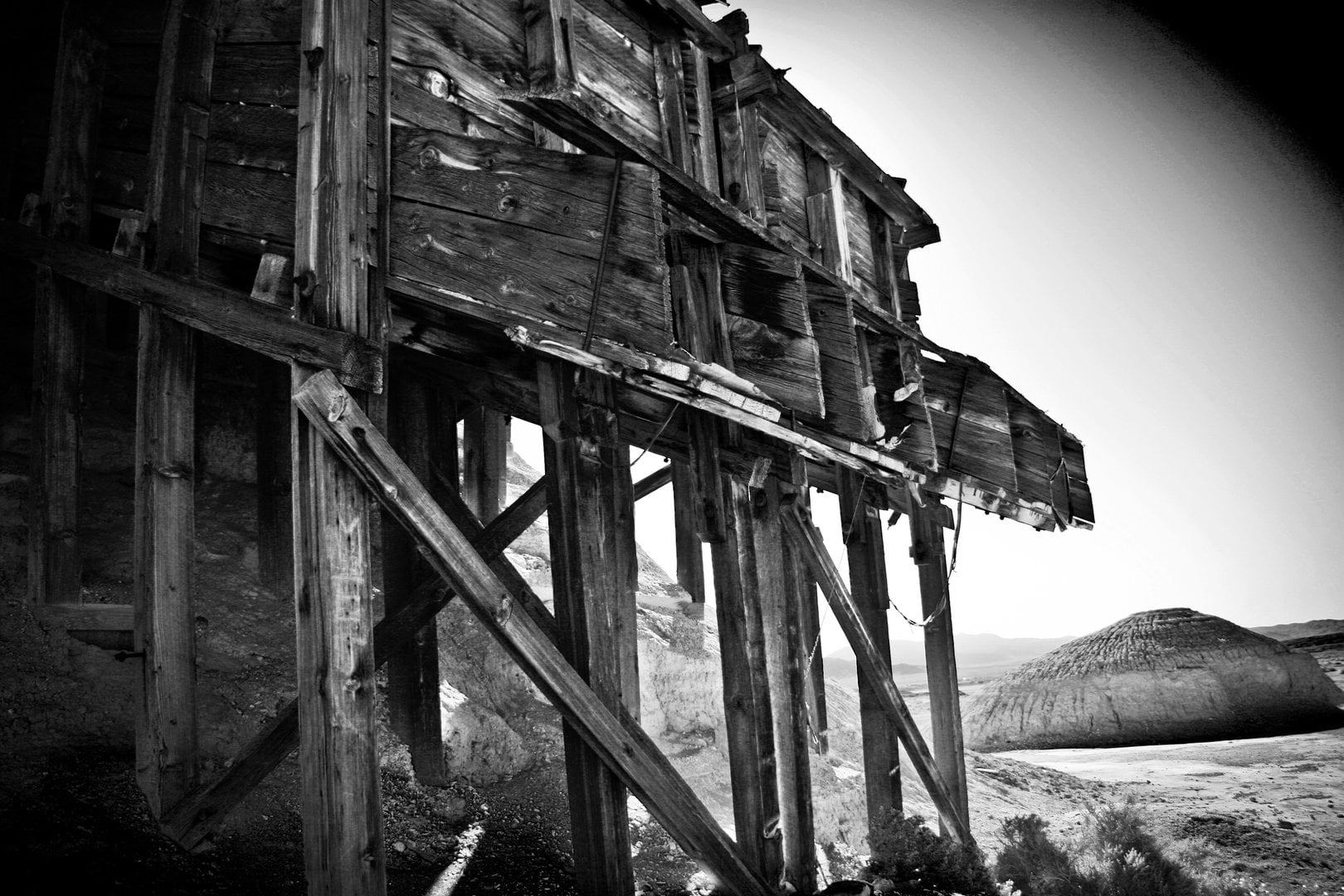 A decaying wooden structure stands against the stark landscape of Death Valley, its weathered beams framing the desert beyond. Captured in black and white, the image echoes the ghosts of the mining era — a reminder of human grit slowly surrendering to time and sand.